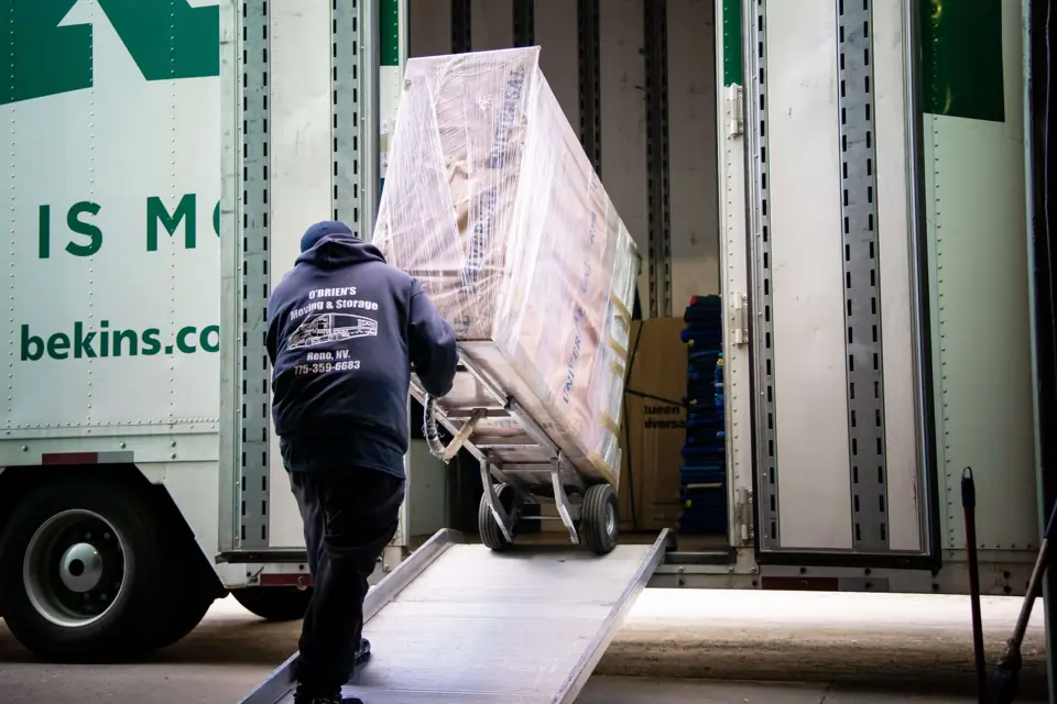 An O'Brien's mover wheeling a shrink-wrapped tall item up the ramp into a Bekins trailer