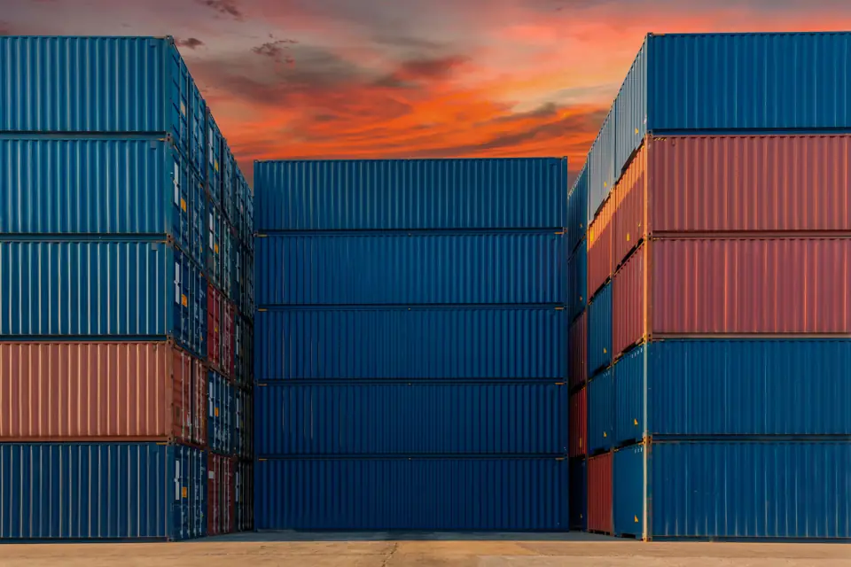 Stacks of blue and red shipping containers at a port under a sunset sky