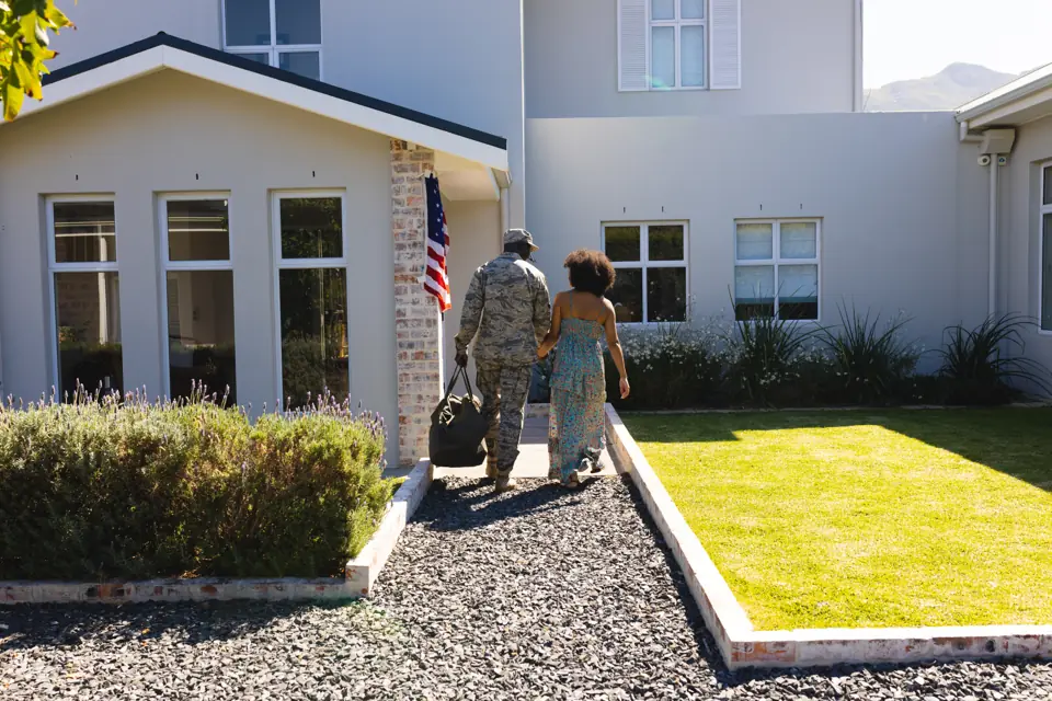 Soldier in camouflage and a woman walking hand in hand toward a house, seen from behind